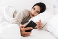 Photo of adorable woman 30s using smartphone, while lying in bed with white linen at home Royalty Free Stock Photo