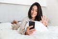Photo of adult woman 30s using mobile phone, while lying in bed with white linen in bright room Royalty Free Stock Photo