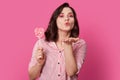 Photo of affectionate woman blows air kiss, dressed in fashionable clothes, holds candy, has bright makeup, poses over rosy