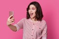 Photo of amazed surprised female holds mobile phone for making selfie portrait, has new image, bright pink makeup, dressed in Royalty Free Stock Photo
