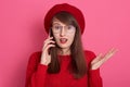 Photo of astonished girl wearing red beret, sweater,spectacles, talking on smartphone with her friend, spreading hand aside, keeps Royalty Free Stock Photo