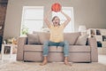 Photo of astonished glad sporty guy sit couch watch basketball match raise ball wear yellow t-shirt jeans home indoors