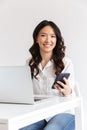 Photo of attractive asian office woman with long dark hair sitting at table and holding smartphone while working with laptop, iso Royalty Free Stock Photo