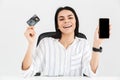 Photo of attractive brunette businesswoman 30s holding smartphone and plastic credit card while sitting in armchair at table in Royalty Free Stock Photo