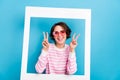 Photo of attractive cheerful female in heart spectacles show v-sign through white frame cadre isolated on blue color Royalty Free Stock Photo