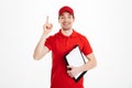 Photo of attractive delivery worker in red t-shirt and cap smiling and pointing finger up on copyspace text or product while hold Royalty Free Stock Photo