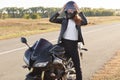 Photo of attractive female biker dressed in leather jacket, wears helmet, poses on motorbike against road background, likes her ho