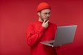 Photo of attractive man with beard in glasses and red clothing. Serious man holds laptop and wonders, over red