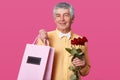 Photo of attractive mature man with pleasant facial expression, dressed in yellow shirt with white bowtie, carries pink bag with