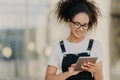 Photo of attractive millennial girl with crisp hair, holds digital tablet, surfes social networks, wears optical glasses, casual t