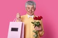 Photo of attractive senior man looks through round spectacles, wears formal shirt and bowtie, holds package with present and red