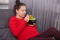 Photo of attractive woman has dark hair bun, going give birth to baby, eats delicious fresh salad, sits at comfortable couch in