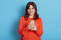 Photo of attractive woman, looks smiling at camera and holding mobile phone, isolated over blue background, winsome lady using