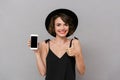 Photo of attractive woman 20s wearing black dress and hat holding smartphone, isolated over gray background