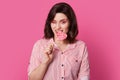 Photo of attractive young woman with dark haired woman with bright makeup, bites candy, blinks eye, poses over pink background, Royalty Free Stock Photo