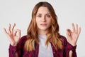 Photo of attractive young woman with dyed hair, makes okay gesture with both hands, confirms somebodys idea, gestures indoor, Royalty Free Stock Photo