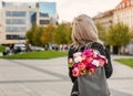 Photo from the back of young blonde romantic woman with bouquet of flowers in grey backpack, standing on a street Royalty Free Stock Photo