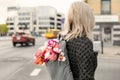 Photo from the back of young blonde romantic woman with bouquet of flowers in grey backpack, standing on a street Royalty Free Stock Photo