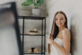 Photo of beautiful European woman combs long hair with hair comb, looks at herself in mirror and smiles gladfully, poses in Royalty Free Stock Photo