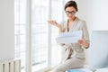 Photo of beautiful female entrepreneur in formal suit, focused in smartphone, checks information from paper documents, holds phone