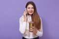 Photo of beautiful girl isolated over lilac background, holding thermo mug in hands, enjoying coffee or tea while talking via Royalty Free Stock Photo