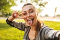 Photo of beautiful sporty woman 20s in sportswear wearing bluetooth earpod taking selfie with fingers, while walking in green park Royalty Free Stock Photo