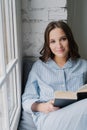 Photo of beautiful woman dressed in casual clothes, wears pyjamas, sits on window sill, reads romantic story in book, enjoys cozin Royalty Free Stock Photo