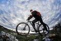 Photo below of the mountain cyclist in the black sportwear on the rocks against dramatic sky with clouds. Royalty Free Stock Photo