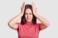 Photo of brunette woman with desperate look, keeps both hands on head, suffers from terrible headache, dressed in casual t shirt,