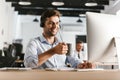 Photo of businessman 30s wearing office clothes and headset, drinking tea from cup while sitting by computer in call center Royalty Free Stock Photo