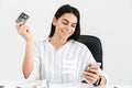 Photo of caucasian brunette businesswoman 30s holding smartphone and plastic credit card while sitting in armchair at table in Royalty Free Stock Photo