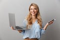 Photo of charming blonde woman 20s dressed in shirt smiling while holding smartphone and silver laptop