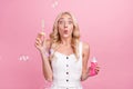 Photo of charming impressed young woman dressed white overall blowing foam bubbles pink color background