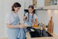 Photo of cheerful busy mum and her daughter pose near stove, serve breakfast for family, fry eggs on frying pan, wear aprons, pose Royalty Free Stock Photo