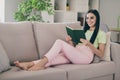 Photo of cheerful clam young lady lay sofa pillows hold book read barefoot wear glasses green t-shirt pink trousers