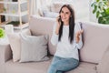 Photo of cheerful positive pretty young lady talk phone raise hand sit living room sofa good mood home indoors Royalty Free Stock Photo