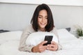 Photo of cheerful woman 30s using mobile phone, while lying in bed with white linen in bright room Royalty Free Stock Photo