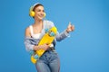 Photo of cheerful young woman with yellow headphones posing with penny or skateboard and showing good gesture over blue