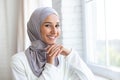 The photo is a close-up portrait of a young beautiful muslim woman in a hijab standing inside by the window, leaning her Royalty Free Stock Photo