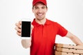 Photo closeup of affable man from delivery service in red t-shirt and cap holding stack of pizza boxes and showing