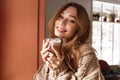 Photo closeup of attractive single woman 20s with brown hair posing on camera, and drinking glass of latte in coffee shop