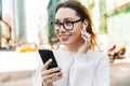 Photo closeup of pretty smiling woman using cellphone and earpod while walking in big city street Royalty Free Stock Photo