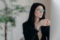 Photo of concentrated brunette female copywriter drinks coffee, works on creative task, wears spectacles and black formal costume