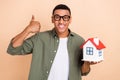 Photo of confident positive man wear khaki shirt spectacles showing thumb up holding small house isolated beige color Royalty Free Stock Photo