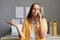 Photo of confused puzzled woman with pleasant appearance sitting in office at her workplace and talking smart phone, shrugging Royalty Free Stock Photo