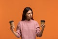 Photo of confused unaware woman in pink sweater chooses between two types of drink in disposable cups.Tea or coffee. Need to cheer