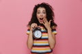 Photo of confused woman 20s with curly hair holding alarm clock, isolated over pink background