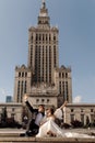 Photo of couple of newlyweds, man and woman, bride inwedding dress man in suit on background of cityscape are sitting on road with