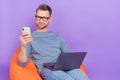 Photo of cute busy guy dressed grey shirt glasses sitting bean bag working gadgets empty space isolated purple color