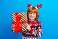 Photo of cute cheerful little boy dressed ugly pullover horns holding x-mas present isolated blue color background Royalty Free Stock Photo